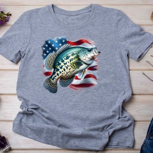Op de afbeelding: Een grijs T-shirt met een afbeelding van een groene en blauwe vis die voor een Amerikaanse vlag zwemt.