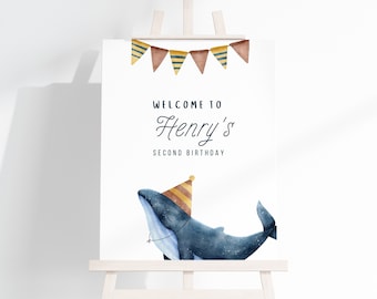 May include: A white welcome sign with a blue whale wearing a yellow and brown party hat. The sign says "Welcome to Henry's Second Birthday" with a string of brown, yellow, and blue triangle flags at the top.