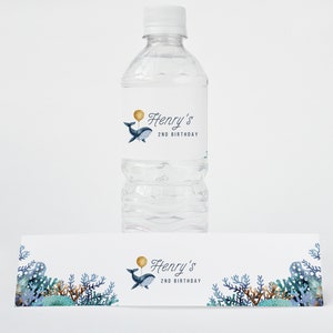 May include: Water bottle label featuring a blue whale holding a gold balloon. The label reads "Henry's 2nd Birthday". A matching banner with a whale and coral reef design is also included.