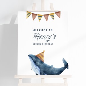 May include: Welcome to Henry's Second Birthday party sign featuring a blue whale wearing a yellow and brown party hat. The sign has a banner of brown, yellow, and blue triangular flags.