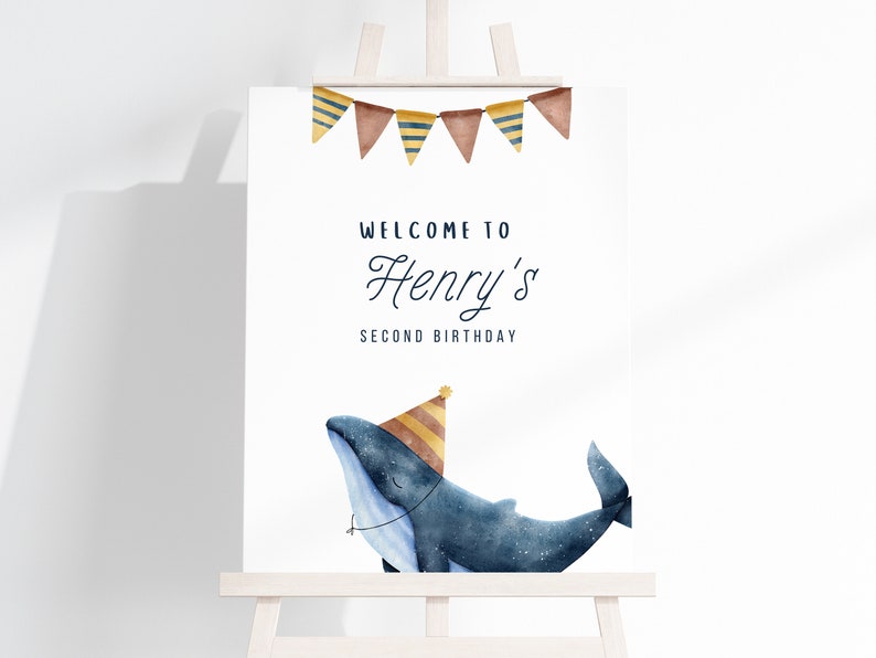 May include: Welcome to Henry's Second Birthday party sign featuring a blue whale wearing a yellow and brown party hat. The sign has a banner of brown, yellow, and blue triangular flags.