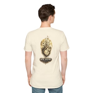 Puede incluir: Una camiseta de manga corta color crema con un gráfico en la espalda. El diseño muestra un corazón anatómico con flores que brotan de la parte superior, frente a una luna. Debajo del corazón, el texto dice NEW HEART EZEKIEL 36:26.