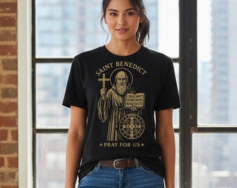 May include: Black t-shirt featuring a gold illustration of Saint Benedict holding a cross and a book. The text on the shirt reads "SAINT BENEDICT" and "+ PRAY FOR US +". The design includes the words "CRUX SANCTI PATRIS PATRIS BENEDICTI".