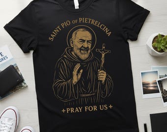 May include: Black t-shirt featuring a gold illustration of Saint Pio of Pietrelcina holding a crucifix. The text "SAINT PIO of PIETRELCINA" arches above the image, with "+ PRAY FOR US +" below. The shirt is a casual wear item.