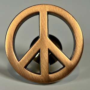 May include: A copper-colored peace sign with a brushed finish. The peace sign is attached to a pin back.