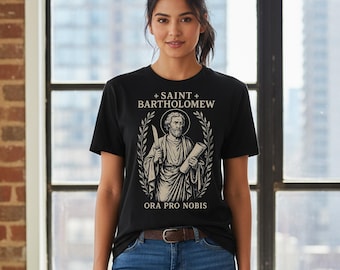 May include: Black t-shirt featuring a graphic of Saint Bartholomew holding a knife and scroll, surrounded by a laurel wreath. The text "SAINT BARTHOLOMEW" and "ORA PRO NOBIS" are printed above and below the image. The model is wearing blue jeans.