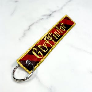 May include: A red and black striped fabric keychain with a gold embroidered "Gryffindor" inscription. The keychain has a silver metal ring.