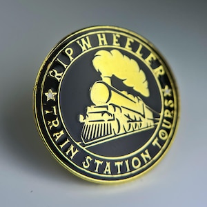 May include: A round, gold-rimmed pin with a black center featuring a gold train illustration. The pin reads "RIP WHEELER TRAIN STATION TOURS" in a circular pattern around the train image. Two gold stars are on the left side.