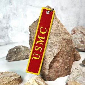 May include: Red fabric keychain with a yellow embroidered "USMC" logo. The keychain is hanging from a silver key ring.