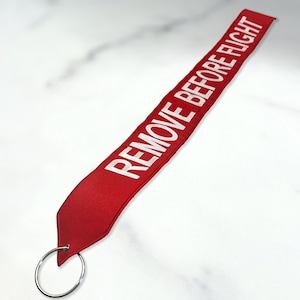 May include: Red fabric keychain with a silver ring and the text "REMOVE BEFORE FLIGHT" in white.