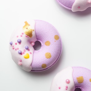 Könnte beinhalten: Draufsicht auf drei dekorative Donuts. Jeder Donut ist zur Hälfte lavendelfarben und zur Hälfte weiß mit Streuseln und goldenen Akzenten. Die Donuts haben ein Loch in der Mitte und sind auf einer weißen Oberfläche angeordnet.