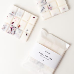 Peut inclure: Trois barres de cire de soja blanches avec un motif floral. L'une est emballée dans du cellophane transparent avec une étiquette indiquant "Rainy Day ROSE & SWEET PEA soy wax melt bar". Les deux autres sont en vrac et ont un motif quadrillé.