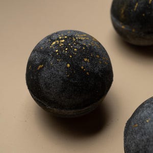 Könnte beinhalten: Nahaufnahme von drei schwarzen Badebomben mit Goldflocken. Die kugelförmigen Bomben haben eine strukturierte Oberfläche und sind auf einem neutralen Hintergrund angeordnet. Die goldenen Akzente verleihen den Badeprodukten einen Hauch von Luxus.