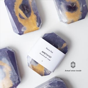 Pode incluir: Várias barras de sabonete artesanal roxo e dourado embrulhadas em plástico transparente. Uma barra apresenta um rótulo de papel branco com o texto Rainy Day AMETHYST handmade soap e um ícone com o texto Actual stone inside.