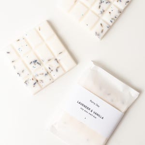May include: Three white soy wax melt bars with lavender and vanilla. One bar is in a paper wrapper with a label that reads "Lavender & Vanilla soy wax melt bar". The bars are divided into squares and have lavender buds.