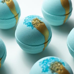 Puede incluir: Varias bombas de baño azul claro con detalles dorados y azules dispuestas sobre una superficie blanca. Cada bomba esférica tiene una franja dorada y un toque de purpurina azul y dorada, evocando un ambiente de spa.