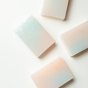 Op de afbeelding: Vier rechthoekige zeepblokken met een kleurengradiënt. Elke zeep verandert van zachtroze naar een lichtblauwe tint. De zepen zijn gerangschikt op een wit oppervlak, wat een strakke en minimalistische uitstraling creëert.