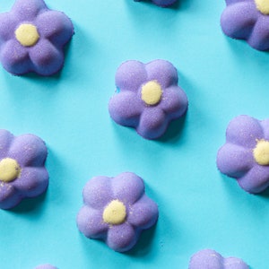 Könnte beinhalten: Ein Muster aus lila, blütenförmigen Badebomben mit gelben Zentren auf einem hellblauen Hintergrund. Die Badebomben haben ein strukturiertes, körniges Aussehen, was auf eine sprudelnde Qualität hindeutet. Die Anordnung ist gleichmäßig verteilt und erzeugt ein ansprechendes Design.