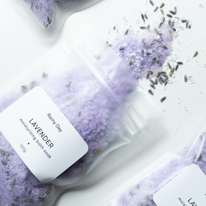 Könnte beinhalten: Klare Plastiktüten gefüllt mit lavendelfarbenen Badesalzen und getrockneten Lavendelblüten. Jede Tüte hat ein weißes Etikett mit "Rainy Day" und "Lavender moisturizing bath soak 100g". Der Hintergrund ist weiß.