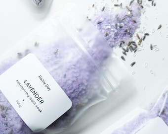 May include: Clear plastic bags filled with lavender-colored bath salts and dried lavender buds. Each bag has a white label that reads "Rainy Day" and "Lavender moisturizing bath soak 100g". The background is white.
