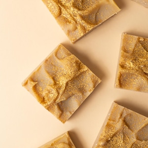 Può includere: Vista dall'alto di diverse saponette quadrate. Il sapone è di colore beige chiaro con accenti dorati. L'oro è a spirale e macchiato sulla superficie, creando un aspetto lussuoso. Le saponette sono disposte su uno sfondo beige chiaro.