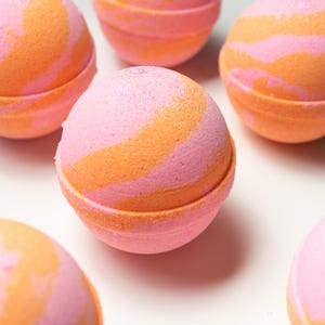 May include: A collection of spherical bath bombs, each featuring a pink and orange marbled pattern. The bath bombs are displayed on a white background. The colour palette consists of bright pink and a warm orange, with a subtle glitter effect.