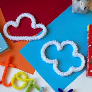 May include: Two fluffy white cloud-shaped decorations on a colourful background. The clouds are made of a soft, textured material. A children's book and a small ceramic animal are also visible. The background is composed of red, blue, and orange paper.
