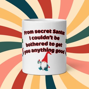 May include: White ceramic mug with the text "From secret Santa; I couldn't be bothered to get you anything good" in red and black. A cartoon gnome with a red hat and a lantern is also featured.