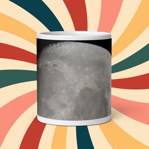 May include: A white ceramic mug with a full-color image of the moon printed on the front. The moon is a close-up of the surface with craters and shadows.