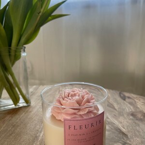 May include: A pink soy wax candle in a glass jar with a pink flower on top. The label reads "FLEURIR | FLO-RIR | FRENCH | (VITO BLOOM. TO BLOSSOM TO FLOURISH | organic soy wax | BLACK RASPBERRY VANILLA"