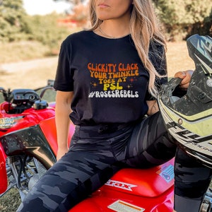 May include: A woman wearing a black t-shirt with the text "Clickity Clack Your Twinkle Toes at PSL #RosesRebels" in orange and yellow lettering. She is sitting on a red ATV and holding a black helmet.