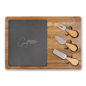 May include: A wooden cheese board with a dark gray slate insert and three cheese knives. The board has the text "Gray Urban Eats" etched on the slate. The knives have wooden handles and stainless steel blades.