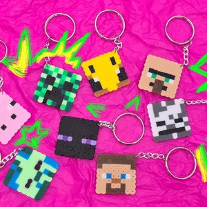 Può includere: Nove portachiavi colorati di personaggi di Minecraft realizzati con perline Perler. I portachiavi presentano personaggi come Steve, Creeper, Enderman e un maiale.