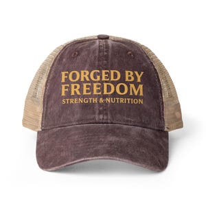 May include: A dark brown distressed trucker hat with a mesh back and tan panels. The front features the gold-colored text FORGED BY FREEDOM STRENGTH & NUTRITION printed in a bold, capitalized serif font across the center of the cap.