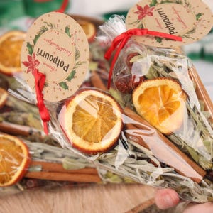 Puede incluir: Primer plano de bolsas de popurrí llenas de rodajas de naranja secas, palitos de canela y hierbas. Cada bolsa está atada con una cinta roja y tiene una etiqueta redonda que dice "Potpourri" con un diseño floral.