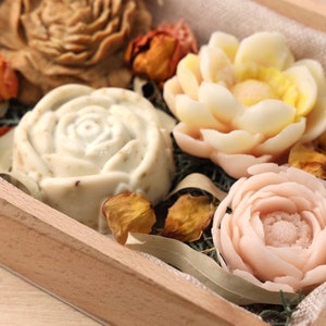 Puede incluir: Una caja de madera llena de cuatro flores de jabón hechas a mano. Los jabones tienen forma de rosas y son de color blanco, beige y rosa. La caja está forrada de arpillera y llena de pétalos de flores secas.