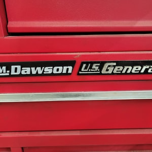May include: Close-up of a red tool chest with the text "C.M. Dawson" and "U.S. General" in silver lettering. The chest has a silver handle and is likely used for storing tools.