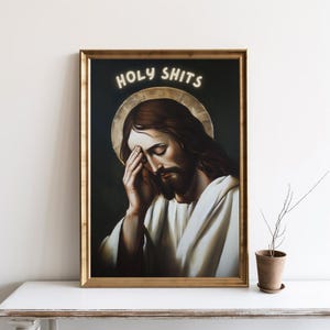 May include: A framed print of a painting depicting Jesus with his head in his hand. The painting is set against a dark background with a halo around Jesus' head. The text "HOLY SHITS" is written in white letters above Jesus' head.