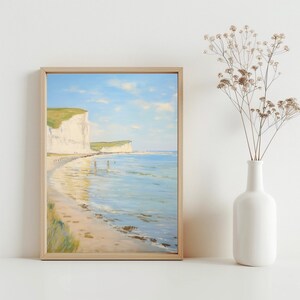 Op de afbeelding: Een ingelijst schilderij van een wit klifstrand met blauw water en een lichtblauwe lucht. Het strand is zanderig met voetafdrukken en het water is helder en kalm.