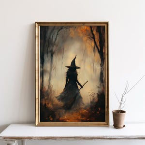 May include: A framed art print featuring a silhouetted witch in a long dress and hat, holding a broom. The artwork is set in a misty forest with a warm, autumnal color palette of browns, oranges, and blacks. The frame is gold.