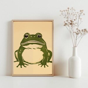 May include: A framed print of a green frog on a light yellow background. The frog is facing the viewer with its mouth slightly open.