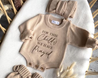 May include: A beige knit baby bodysuit with long sleeves and ribbed cuffs, featuring the embroidered text "FOR THIS Child WE HAVE Prayed" with a small heart. The set includes a matching knit headband and a pair of knit baby booties.