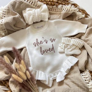 Pode incluir: Um conjunto de roupa de bebé creme, incluindo um body de manga comprida com o texto bordado "she's so loved", uma faixa de cabeça a condizer e botinhas. O conjunto é exibido numa manta de tons neutros com flores secas.