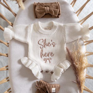 Peut inclure: Une barboteuse à manches longues en tricot côtelé crème avec le texte brodé "She's here" et un petit nœud, accompagnée d'un bandeau noué marron assorti et d'une paire de chaussons en tricot marron avec des liens et une petite étiquette.