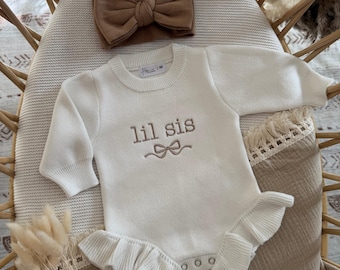 May include: A cream-colored baby bodysuit with the embroidered text "lil sis" and a matching bow headband. The bodysuit has long sleeves and a ruffled skirt. The bow and thread are a caramel color.