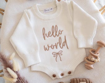 May include: A cream-colored long-sleeve knit romper featuring the embroidered text hello world and a small bow graphic. The romper has brown buttons at the shoulder and bottom. Included are a matching brown knit headband and brown knit booties.