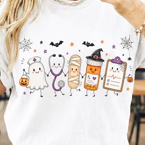 May include: A white t-shirt featuring a graphic of five anthropomorphic medical items holding hands: a ghost nurse with a pumpkin, a stethoscope, a bandage, a prescription bottle with a witch hat, and a clipboard, surrounded by bats and webs.