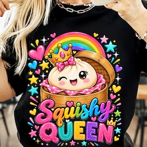 Może przedstawiać: Czarny t-shirt z grafiką puszczającego oko pierożka w koronie, umieszczonego w bambusowym koszyku do gotowania na parze. Wzór zawiera kolorową tęczę, gwiazdki i serca. Napis "Squishy QUEEN" wykonany jest dużymi, wielokolorowymi literami.