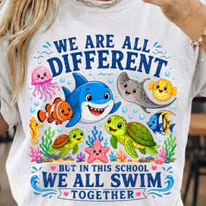 以下が含まれることがあります： サメ、ウミガメ、クラゲ、エイ、魚など、さまざまな海の生き物が描かれたカラフルなグラフィックの白いTシャツ。「WE ARE ALL DIFFERENT BUT IN THIS SCHOOL WE ALL SWIM TOGETHER」という青い文字がプリントされています。