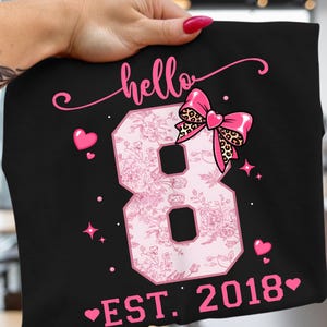 Könnte beinhalten: Ein schwarzes T-Shirt mit einer großen rosa Zahl 8 mit Blumenmuster. Das Design enthält das Wort hello in Schreibschrift, eine rosa Schleife mit Leopardenmuster-Akzent, kleine rosa Herzen und den Text EST. 2018 mit Herzsymbolen auf beiden Seiten.